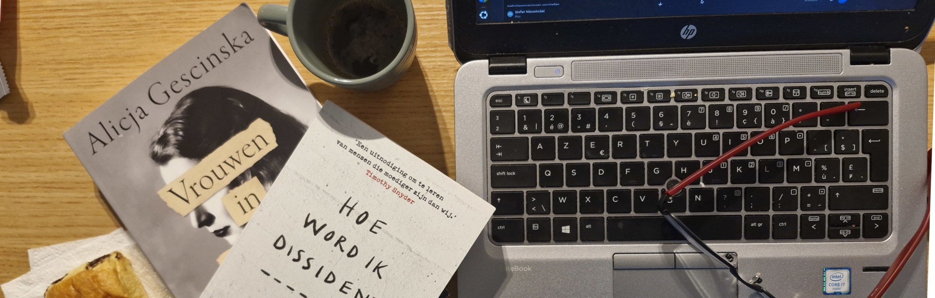 Een schrijftafel met boeken, koffie, laptop en een rode bril